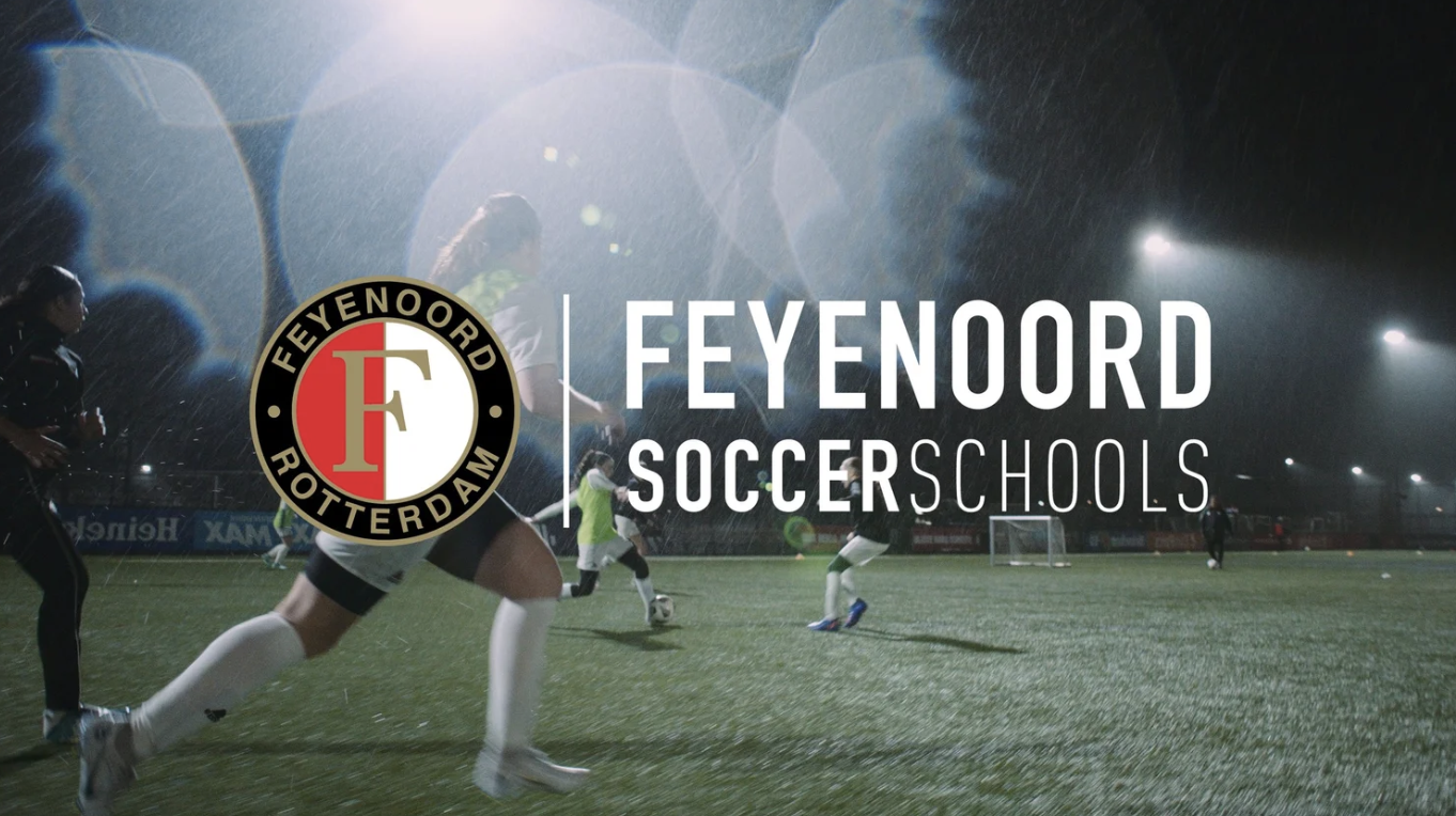 Feyenoord Soccer Schools logo over a soccer game played at night in the rain.