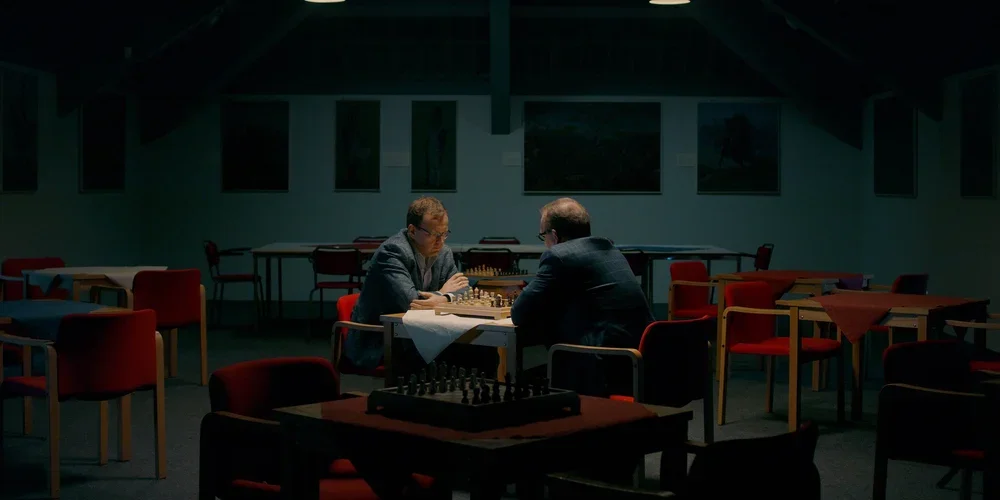 Two men playing chess in a dimly lit, mostly empty room with several tables and red chairs.