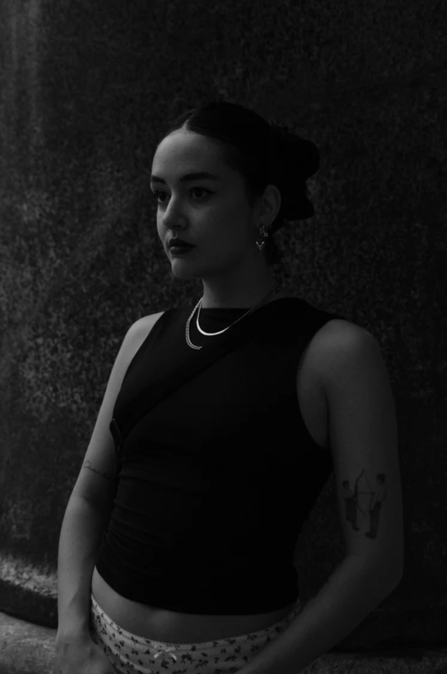A young woman with dark hair styled in a bun, wearing a sleeveless black top, layered necklaces, earrings, and patterned pants, standing against a textured dark wall.