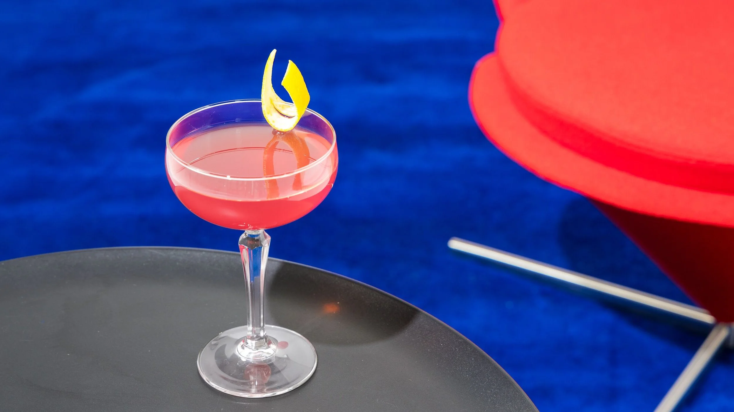 A pink cocktail with a lemon peel garnish in a coupe glass on a black table, with a red lounge chair and blue textured background behind it.