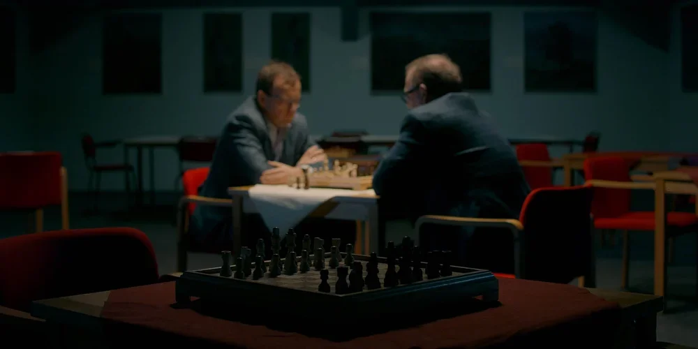 Two men playing chess in a dimly lit room with empty chairs and tables around them and an empty chessboard in the foreground.