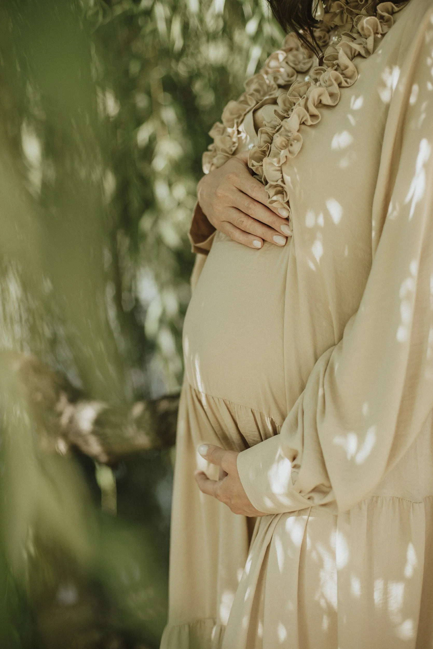 A person wearing a beige dress with ruffled details, holding their pregnant belly outdoors, surrounded by green foliage and dappled sunlight.