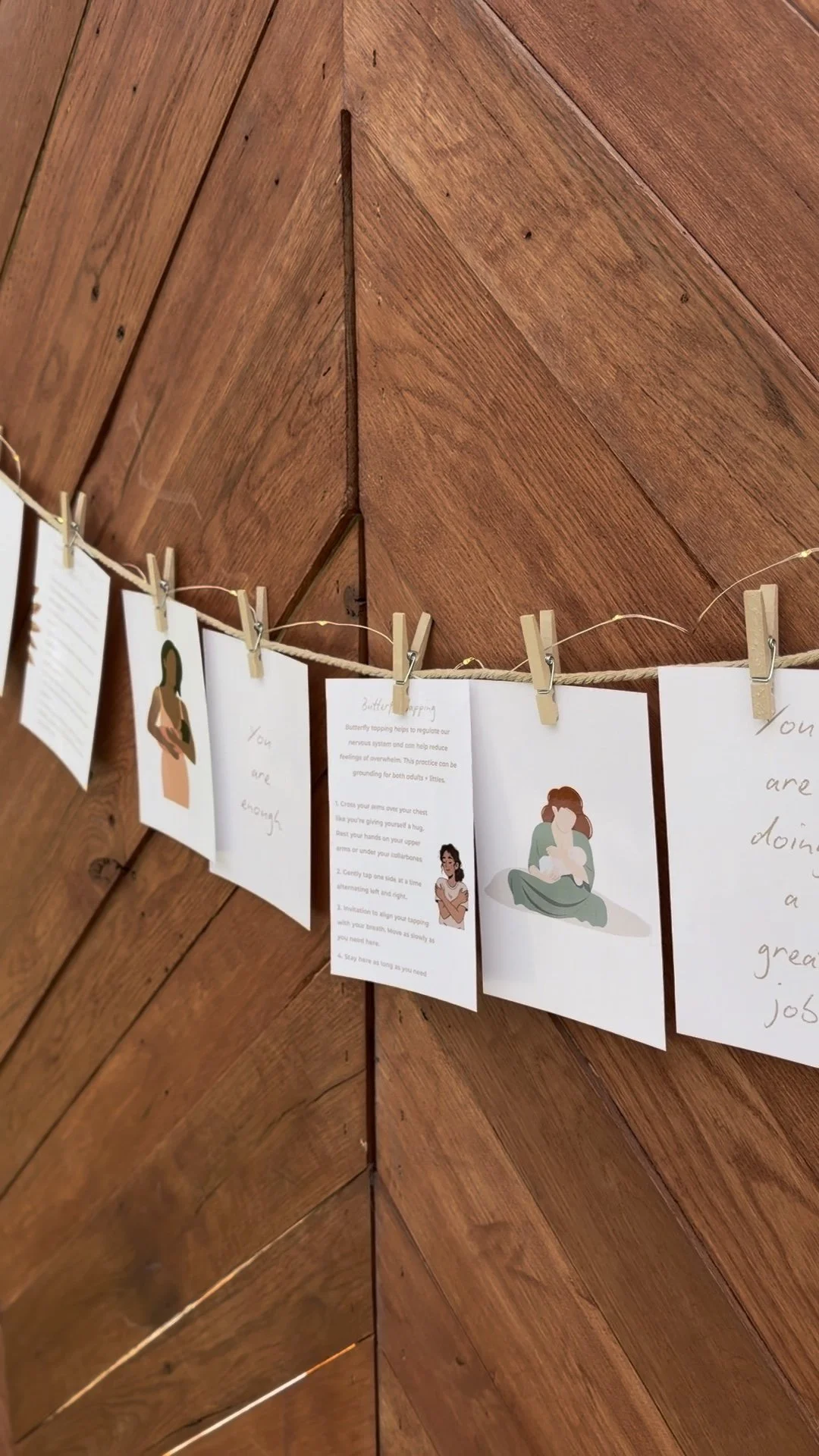 Line of cards with illustrations and handwritten text, clipped to a string with clothespins, hanging against a wooden wall.