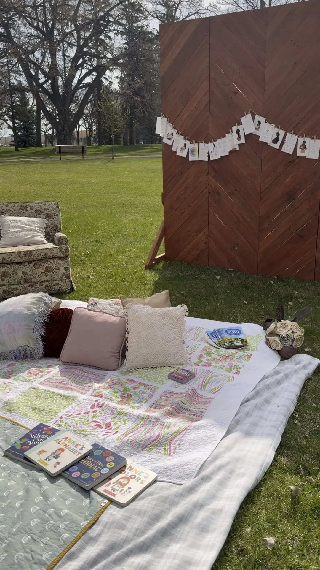 Outdoor reading nook with a white quilt and pillows, books, and a bouquet of flowers on a blanket in a grassy park with trees and a bench in the background.