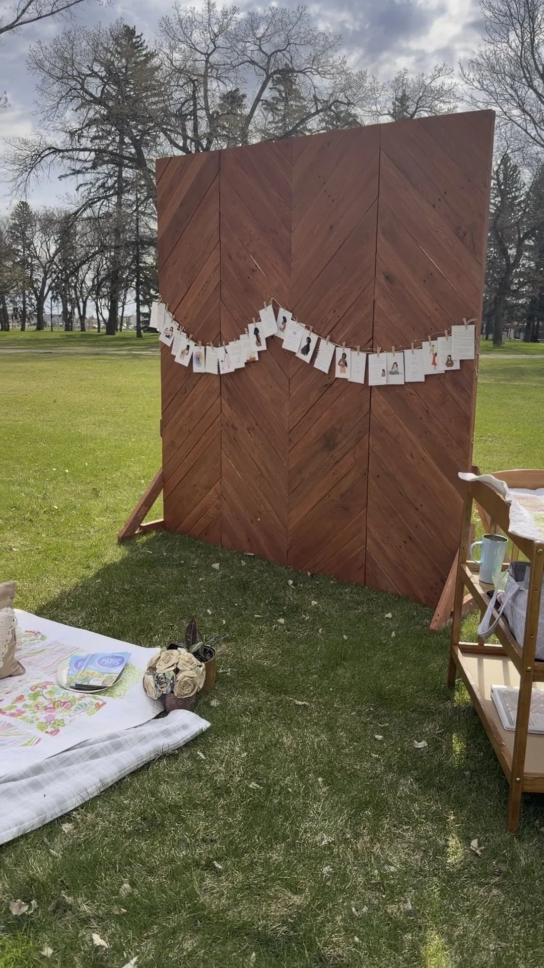 A wooden outdoor display panel with hanging photographs or artwork, set up on a grassy area with trees in the background, during daytime.