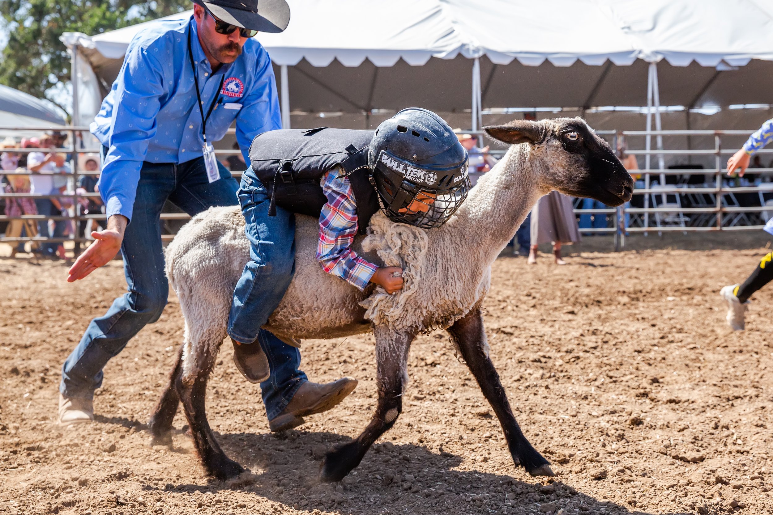 A young boy wearing a helmet, plaid shirt, and vest is riding a sheep during a sheep riding event, with a man assisting and a crowd watching in the background.