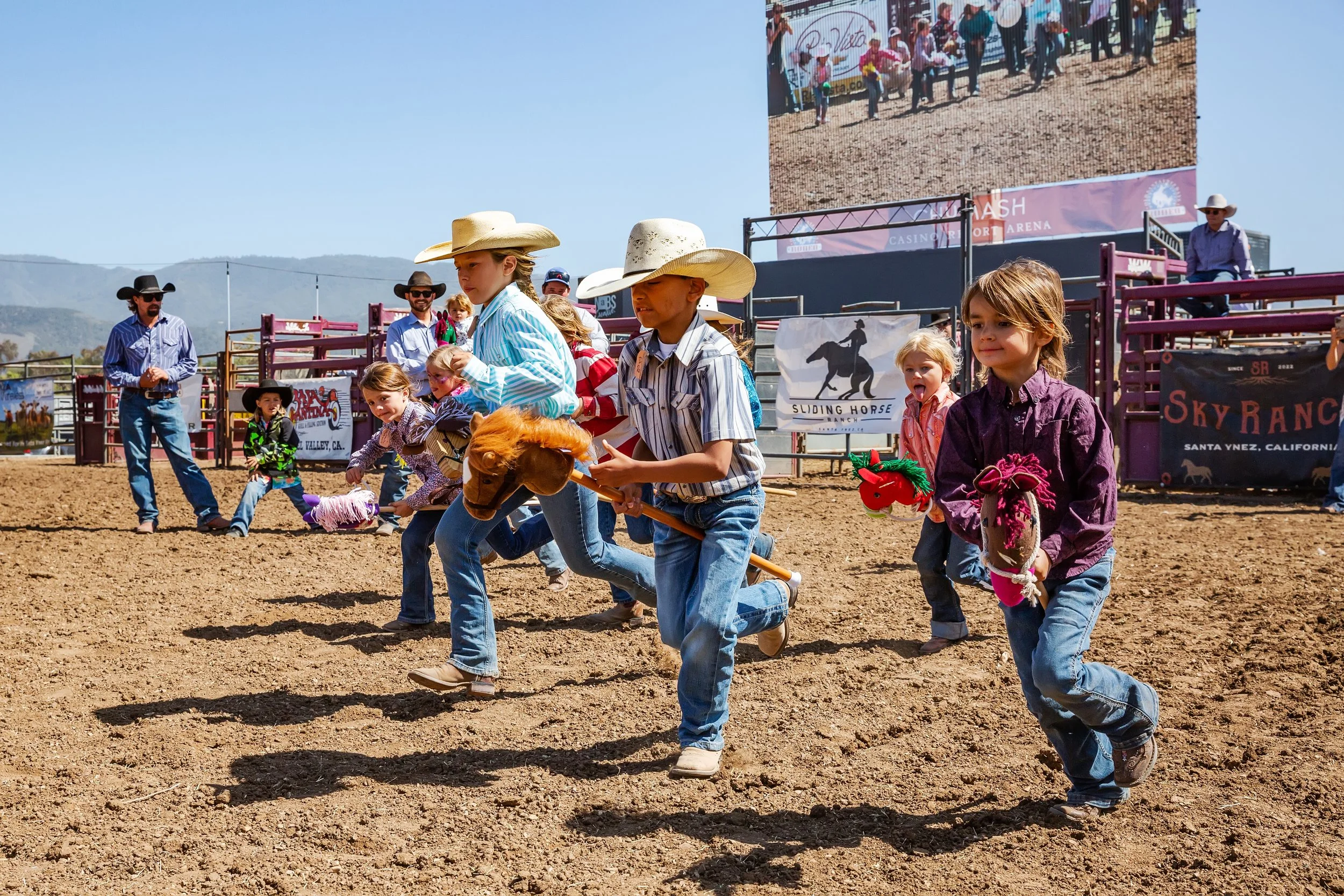 Kids wearing cowboy hats participate in a toy horse race at a rodeo event, with spectators and banners in the background.