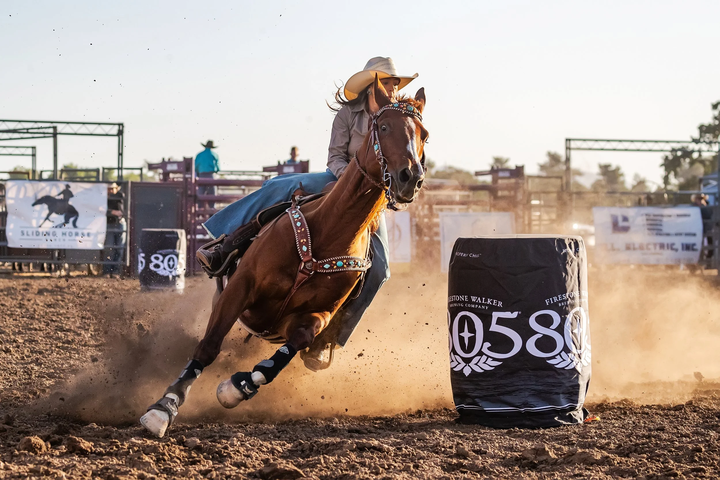 A rodeo rider on a brown horse navigating around a barrel marked with the number '0580', with dust and dirt flying behind. The rider wears a cowboy hat and western attire, during a rodeo event.