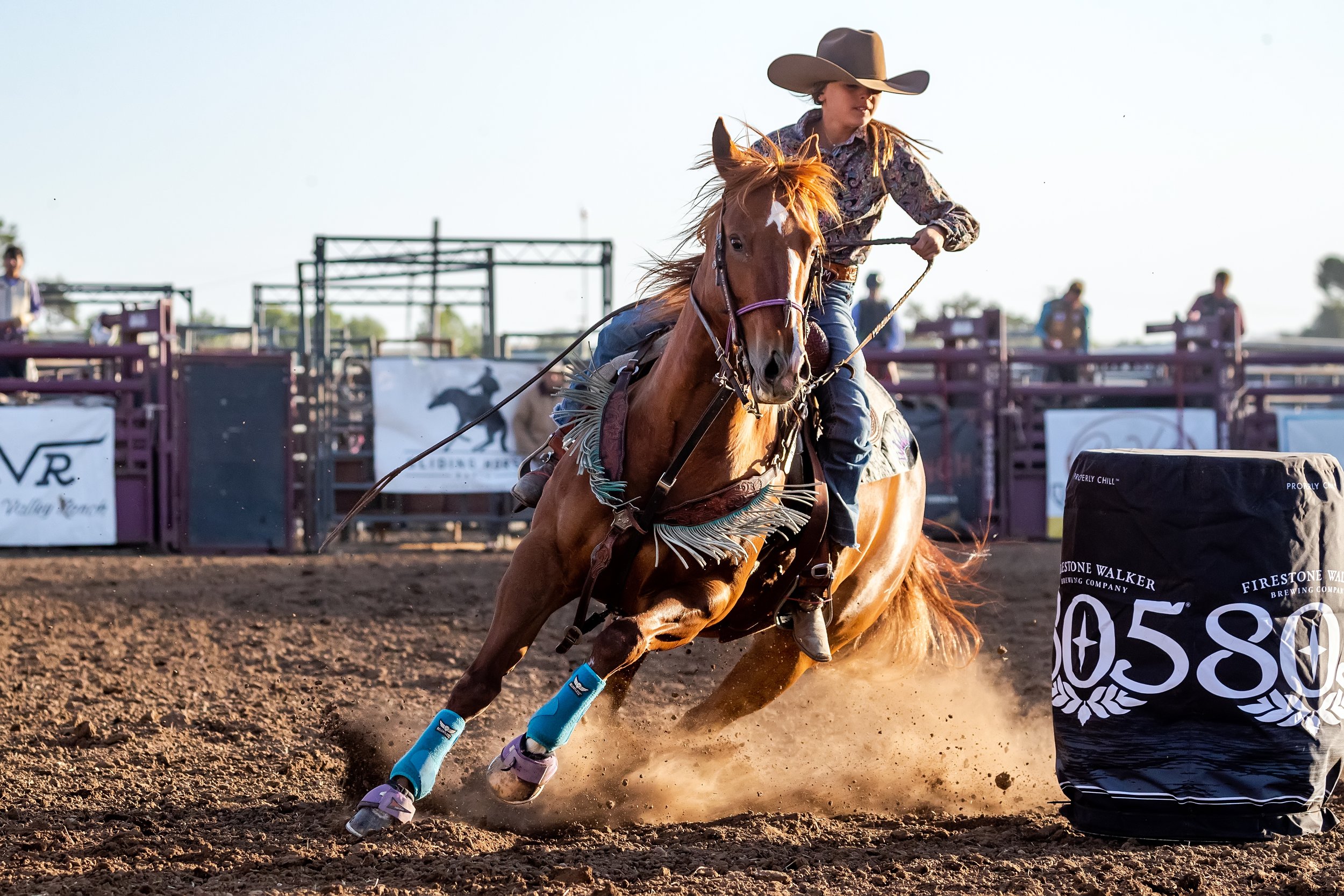 A female cowboy riding a galloping horse around a barrel in a rodeo arena during the daytime.