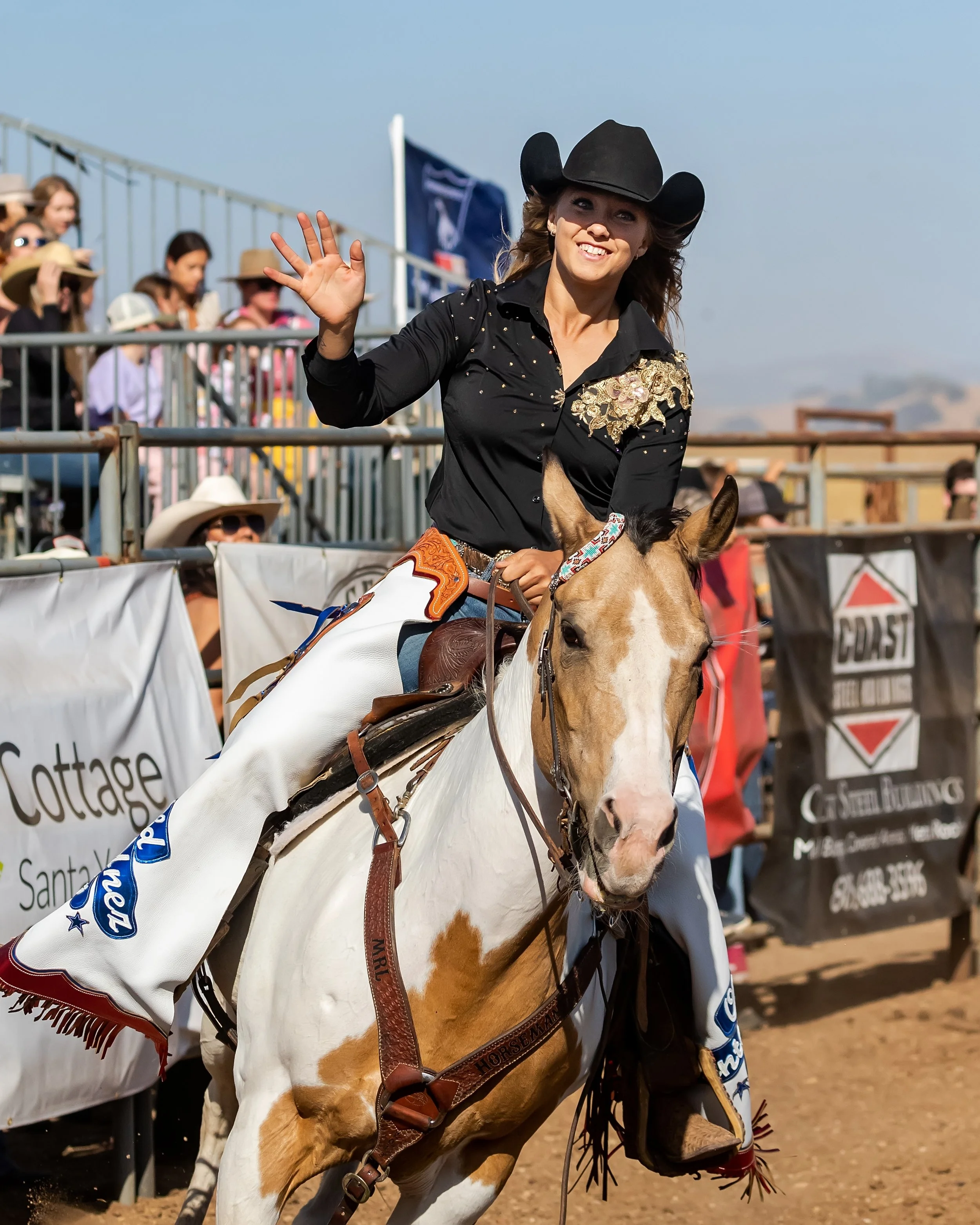 Woman in Western attire riding a paint horse and waving at the camera during a rodeo event with spectators in the background.