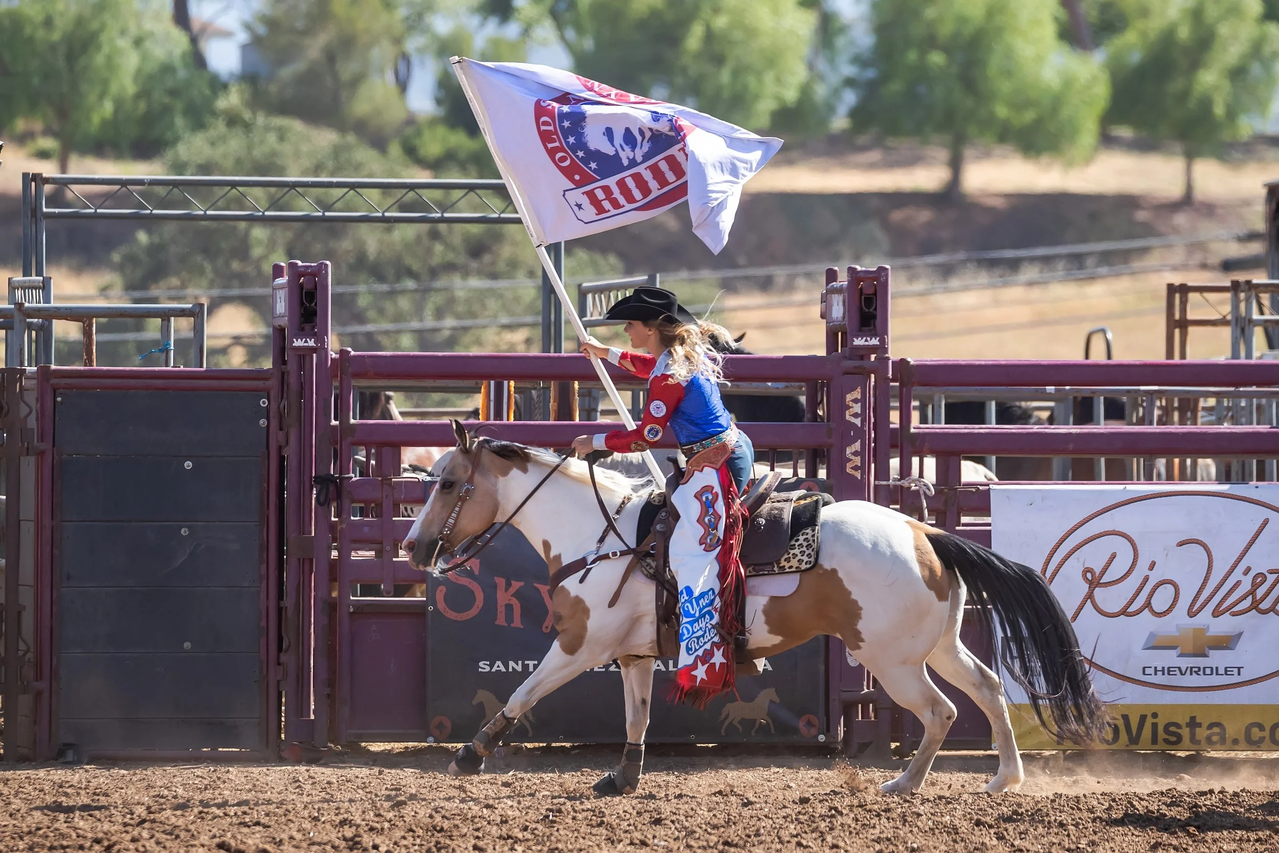 A woman riding a horse during a rodeo event, holding a flag with the rodeo logo, in front of a starting gate at the rodeo arena, with trees and banners in the background.