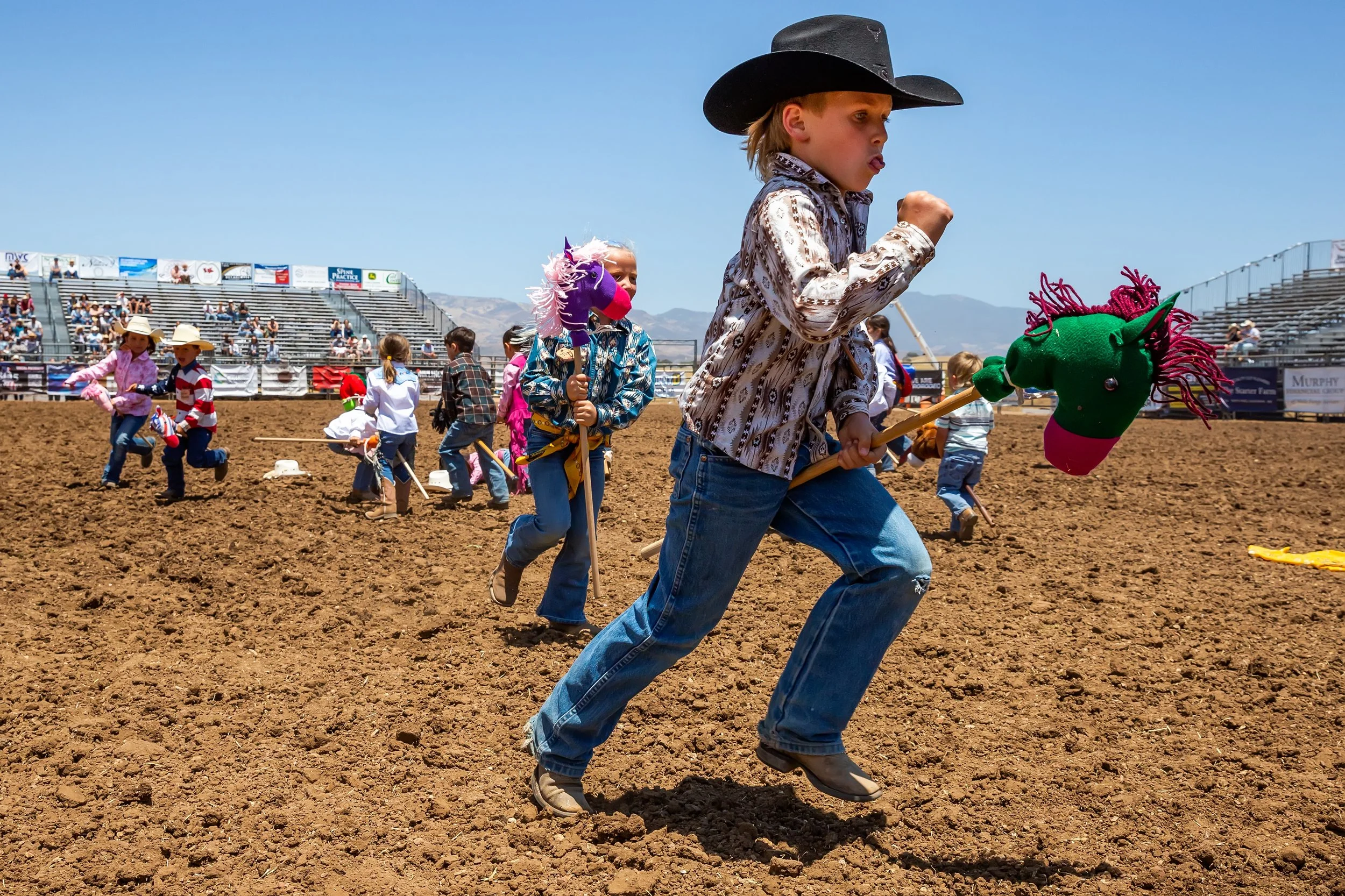 Children participating in a rodeo event, riding hobby horses on a dirt arena, dressed in Western-themed clothing, with some wearing cowboy hats and masks. Spectator stands are visible in the background under a clear blue sky.