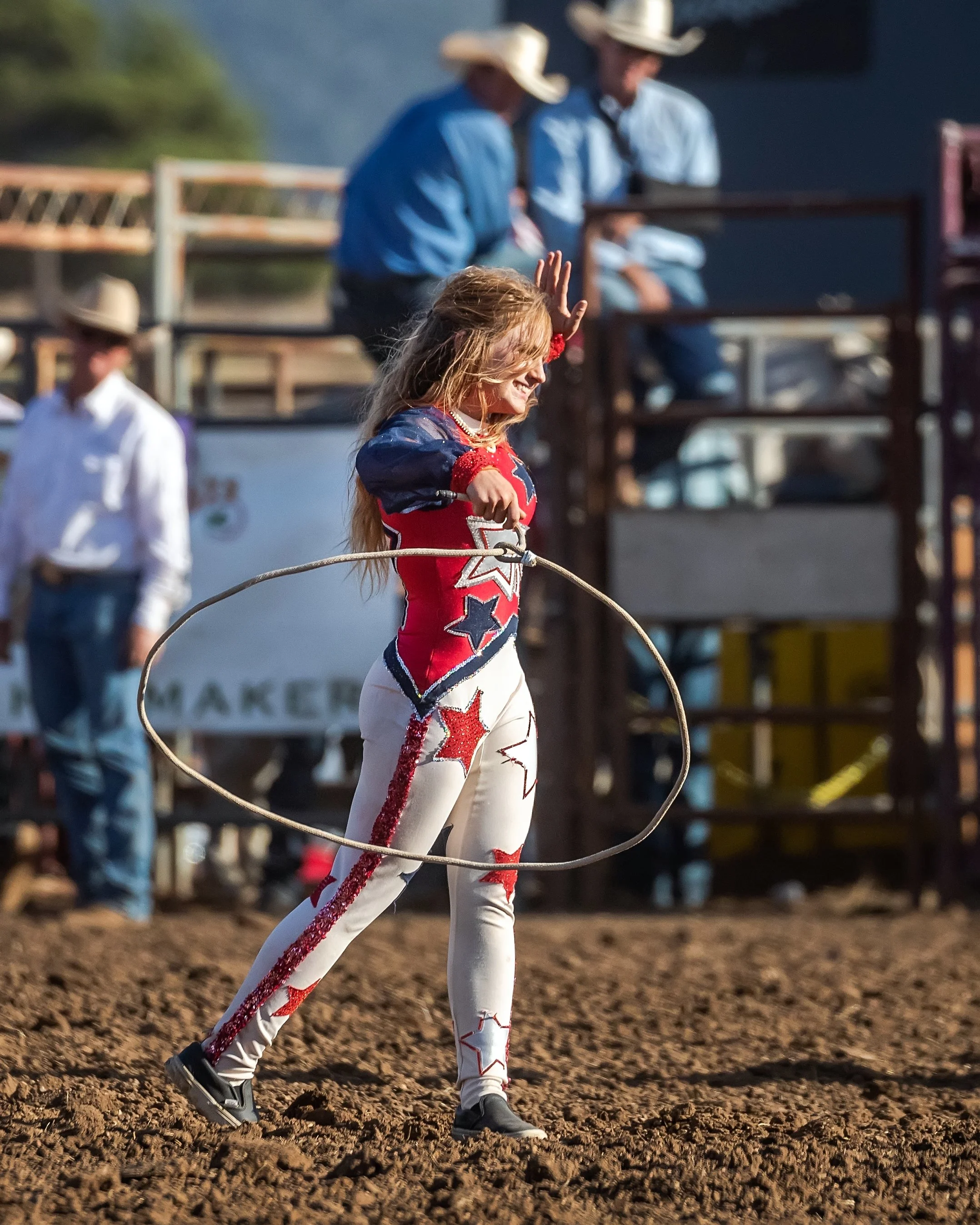 A young girl in a red, white, and blue cowgirl outfit with star patterns, twirling a rope at a rodeo, with people in cowboy hats and western attire in the background.