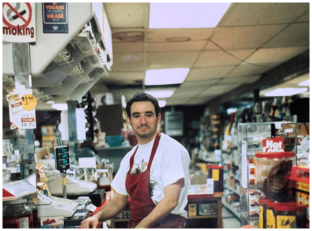 A man working behind the counter at a grocery or convenience store, wearing a white T-shirt and red apron, surrounded by shelves and items for sale.