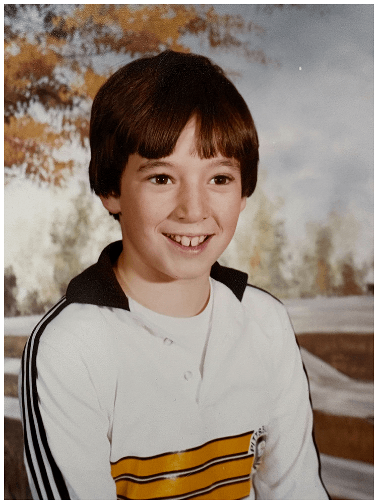 A young boy with brown hair and a big smile, wearing a white and yellow striped long sleeve shirt, standing outdoors with autumn leaves in the background.