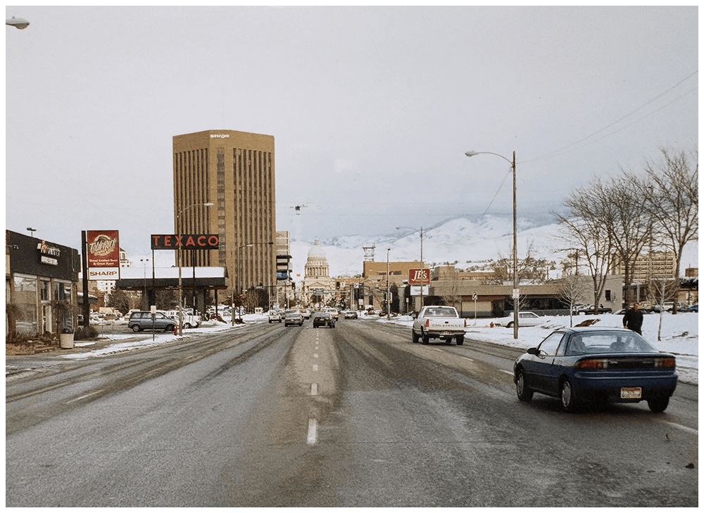 A city street with cars, retail stores, a skyscraper, and mountains in the background on a snowy day.