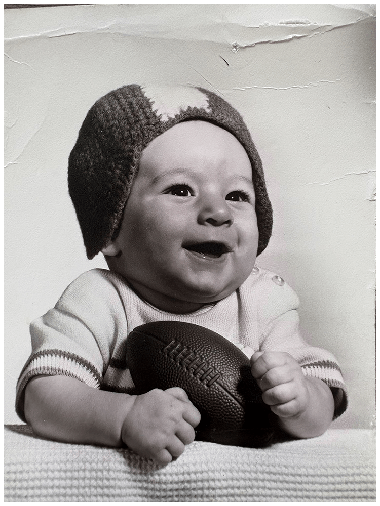 A smiling baby wearing a knit cap and a short-sleeved shirt, holding an American football with both hands, sitting in front of a textured wall.