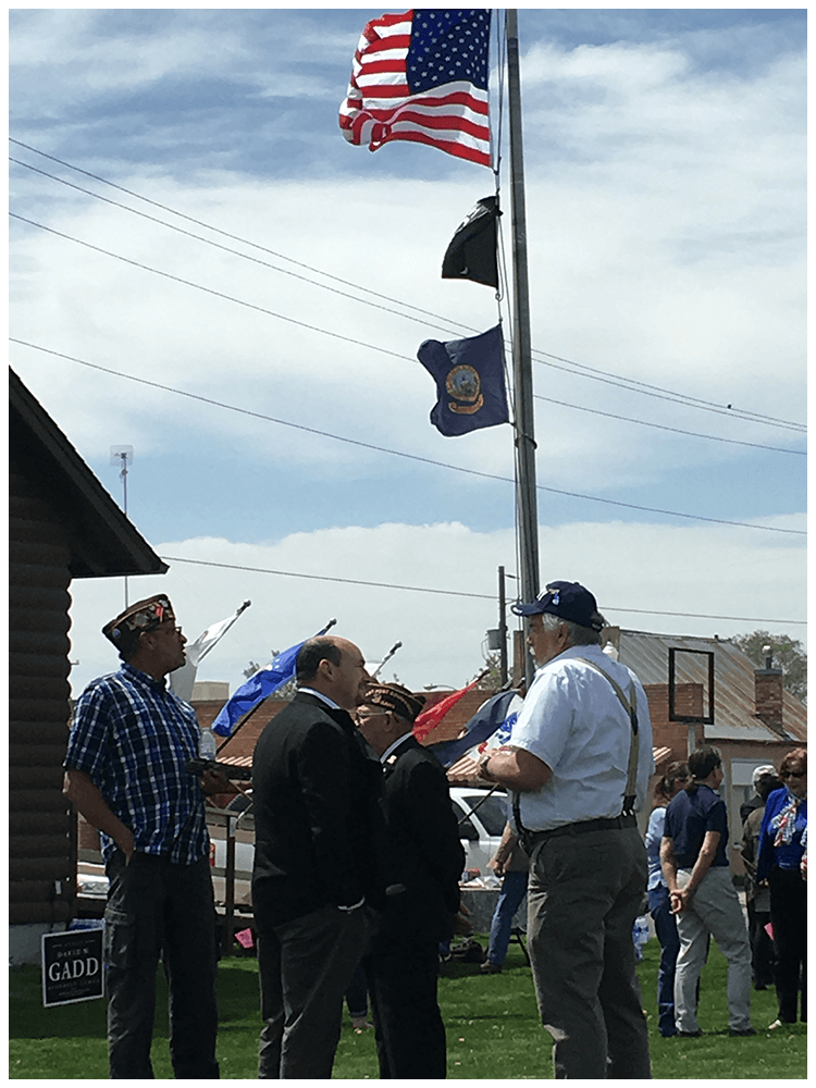 A group of people, including military veterans, are gathered outdoors at a patriotic event with American and military flags flying on a flagpole. Some individuals are wearing military hats, and others are dressed formally, engaging in conversation.