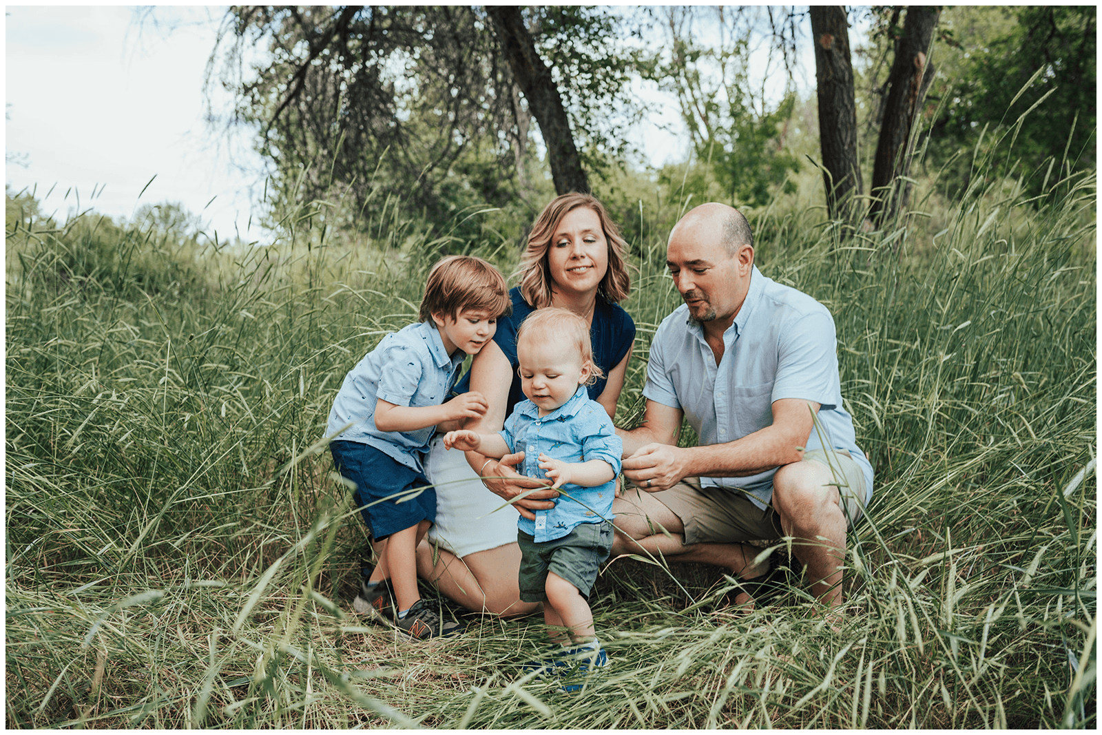 A family of four sitting on the grass in a forested area, playing with a handheld device.