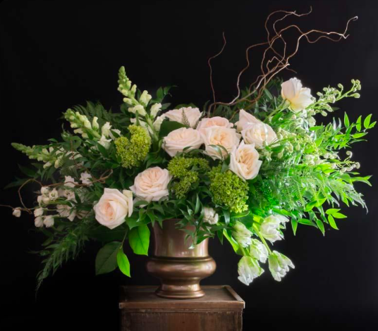 Arrangement of white roses, green hydrangeas, and assorted greenery in a bronze vase on a wooden pedestal against a black background.