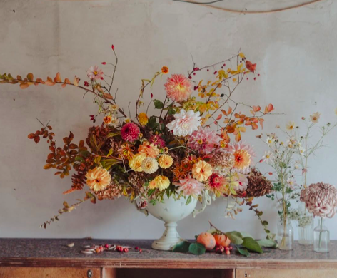 A floral arrangement in a white vase with pink, peach, and cream flowers, placed on a wooden surface with additional flowers and peaches nearby.