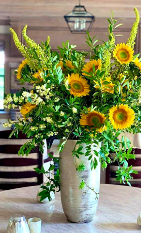 A large bouquet of sunflowers, greenery, and white flowers in a tall white vase on a table.