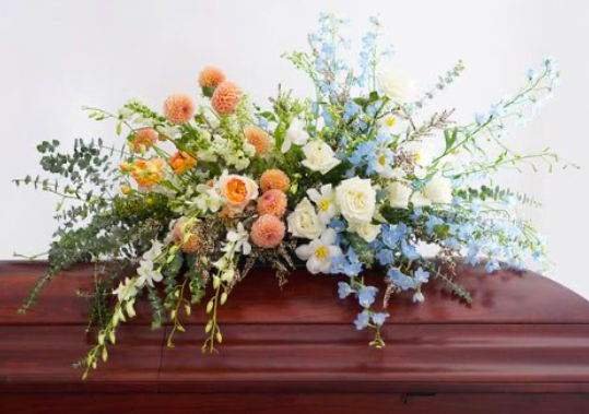 A wooden casket decorated with a large floral arrangement of white, pink, orange, and blue flowers with green foliage.