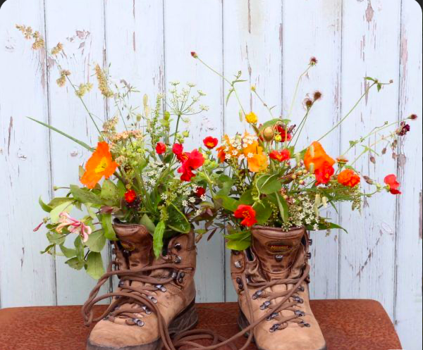 A vase of wildflowers placed inside a pair of worn brown hiking boots on a wooden surface against a white wooden wall.