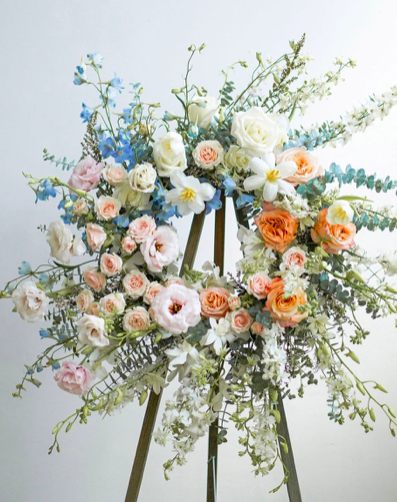 A large floral arrangement on an easel with white, pink, and orange roses, white lilies, blue flowers, and green foliage.