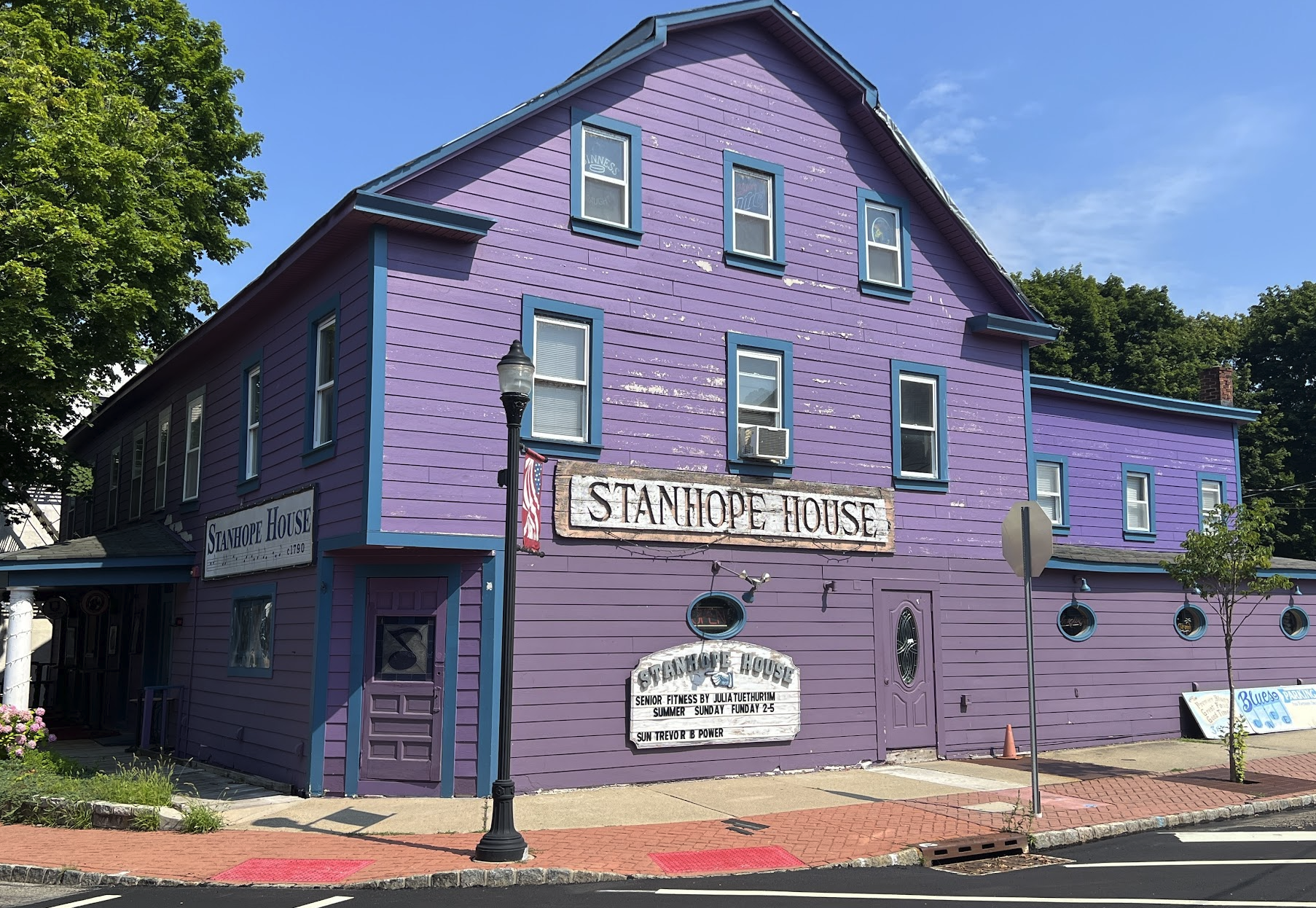 A purple wooden building named Stanhope House with blue trim, multiple windows, and signs indicating it's a senior fitness center. There is a black lamppost, a small tree, and a sidewalk in front of the building, with a clear blue sky overhead.
