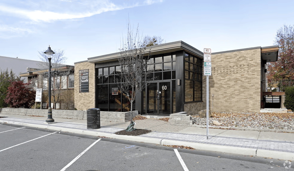 Modern single-story commercial building with brick exterior, large glass windows, and the number 60 on the door, located in a paved parking lot with street parking, a lamppost, and a no parking sign.