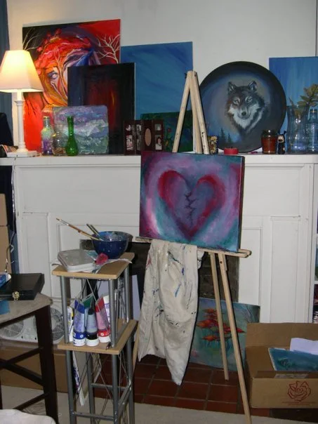 An art studio with paintings on the wall including a portrait of a wolf, abstract art, and a heart with a crack. An easel holds a canvas with a heart and crack design, surrounded by art supplies, bottles, and a lamp.
