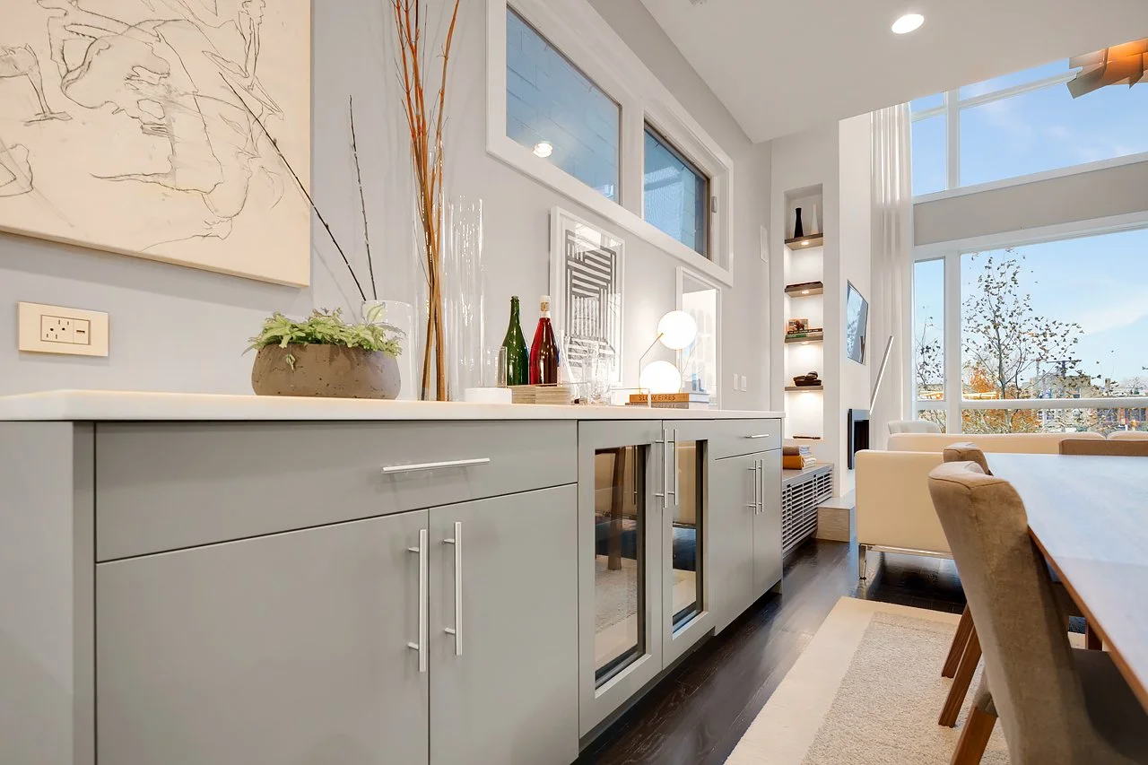 Interior view of a modern living room with large windows, white walls, and a dining table with beige chairs. There is a grey sideboard with decorative bottles, a lamp, and some plants and art on the wall.