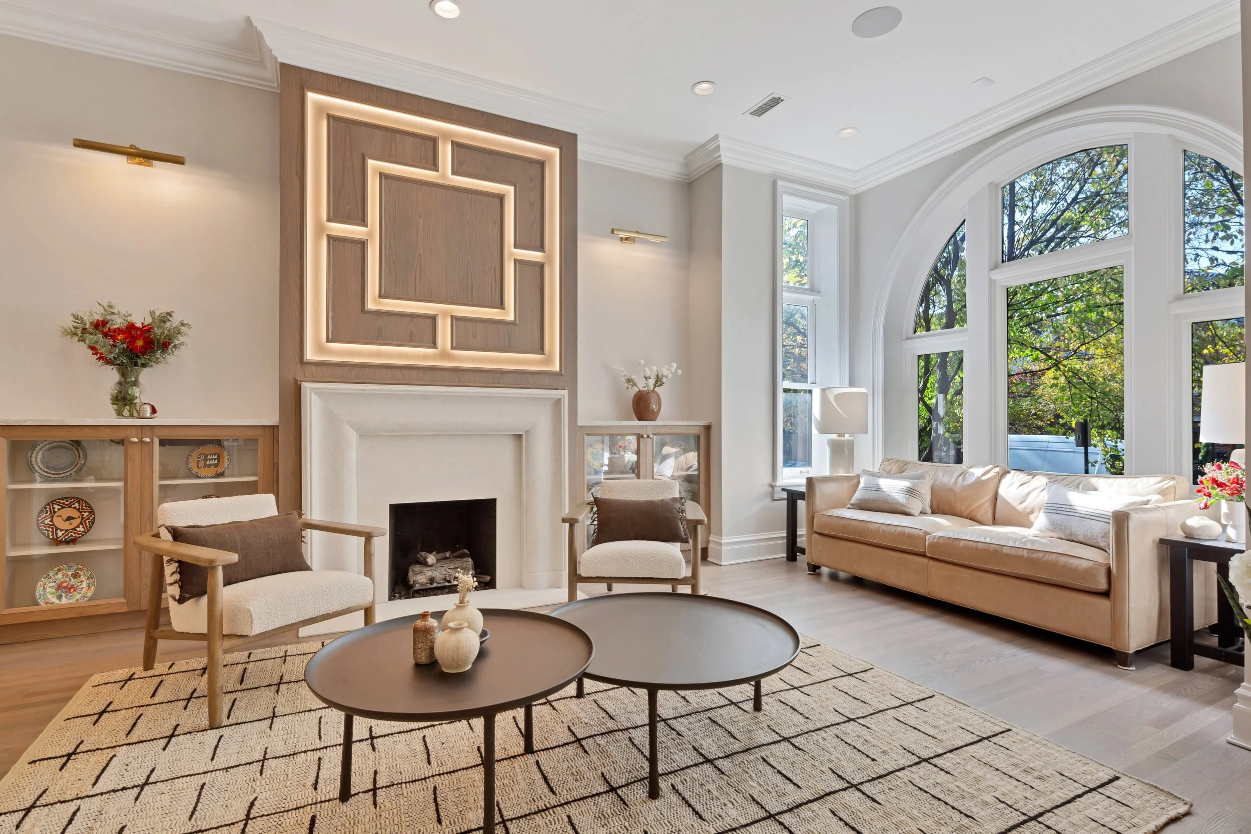 Bright living room with large bay windows, beige sofa, two armchairs, a fireplace, and decorative items on coffee tables and shelves.