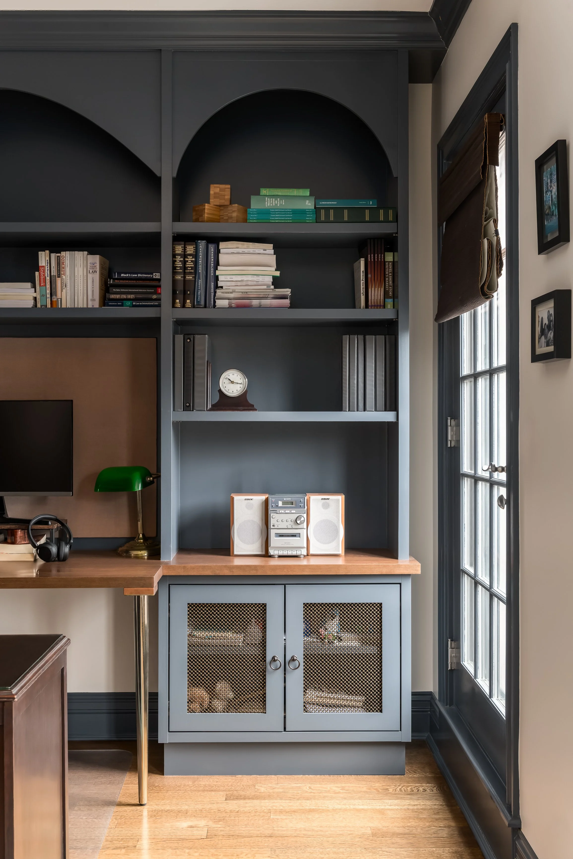 A built-in bookshelf and cabinet in a home office or study, with decorative books, a clock, a stereo system, and a computer monitor, next to a window with blinds.