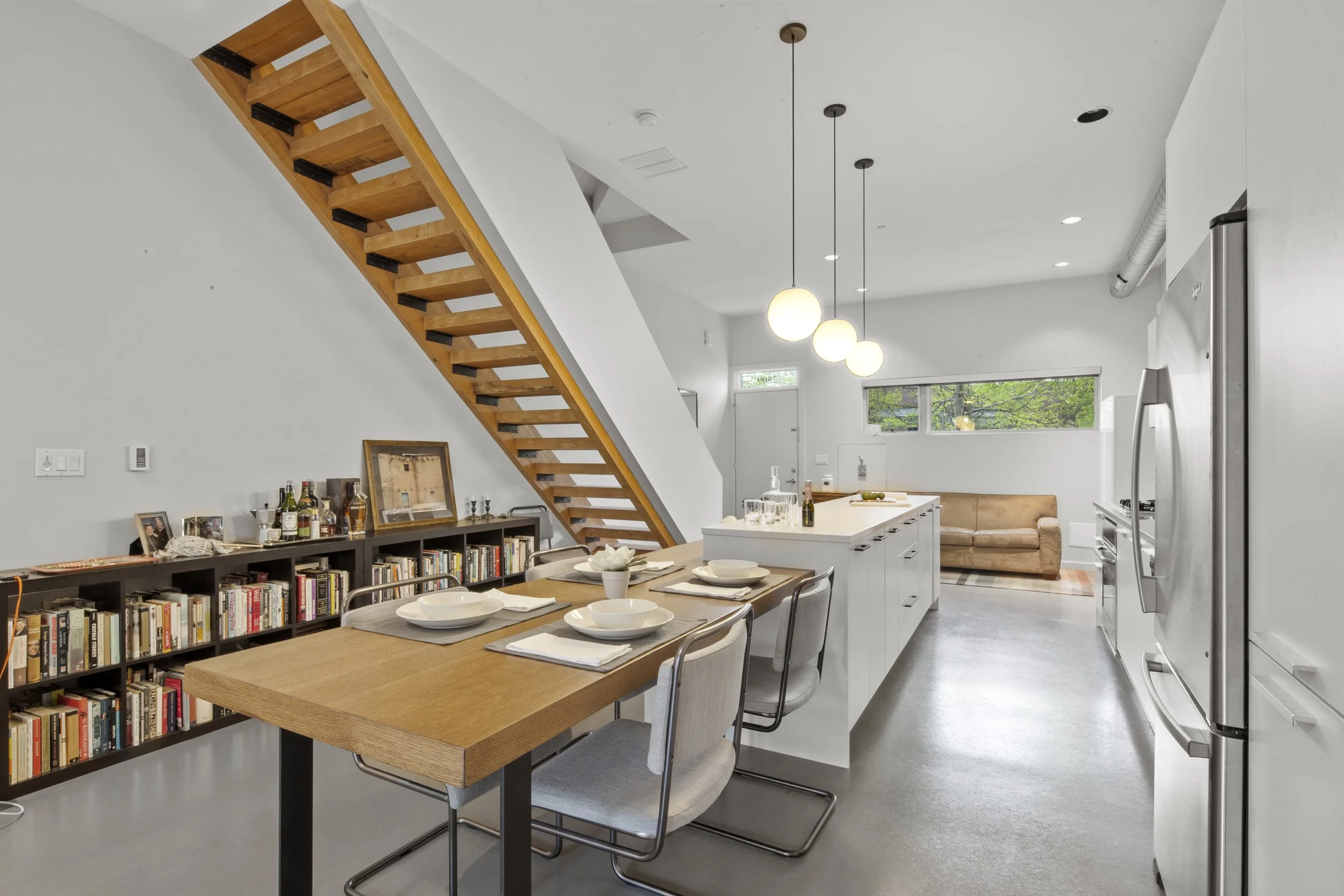 Interior of a modern open-concept kitchen and dining area featuring a wooden dining table with four chairs, white kitchen island with barware, a brown sofa, open shelving with books and bottles, wooden staircase with black supports, and white walls w