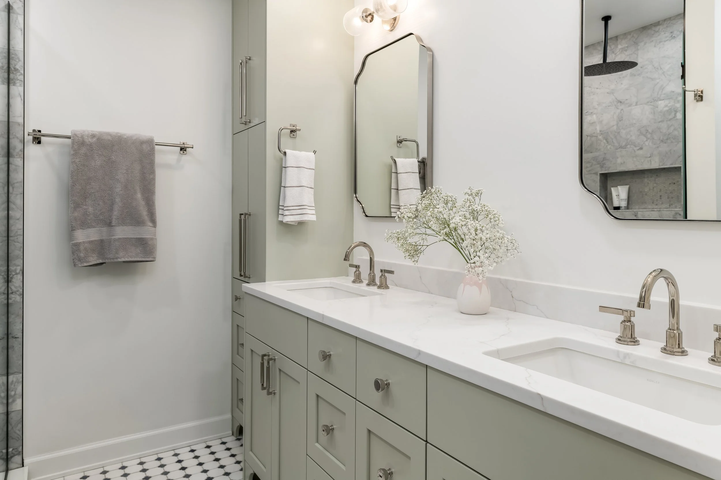 Modern bathroom with double vanity sinks, white marble countertop, two mirrors, wall-mounted lights, gray cabinets, decorative vase with white flowers, and a walk-in shower with marble tiles.