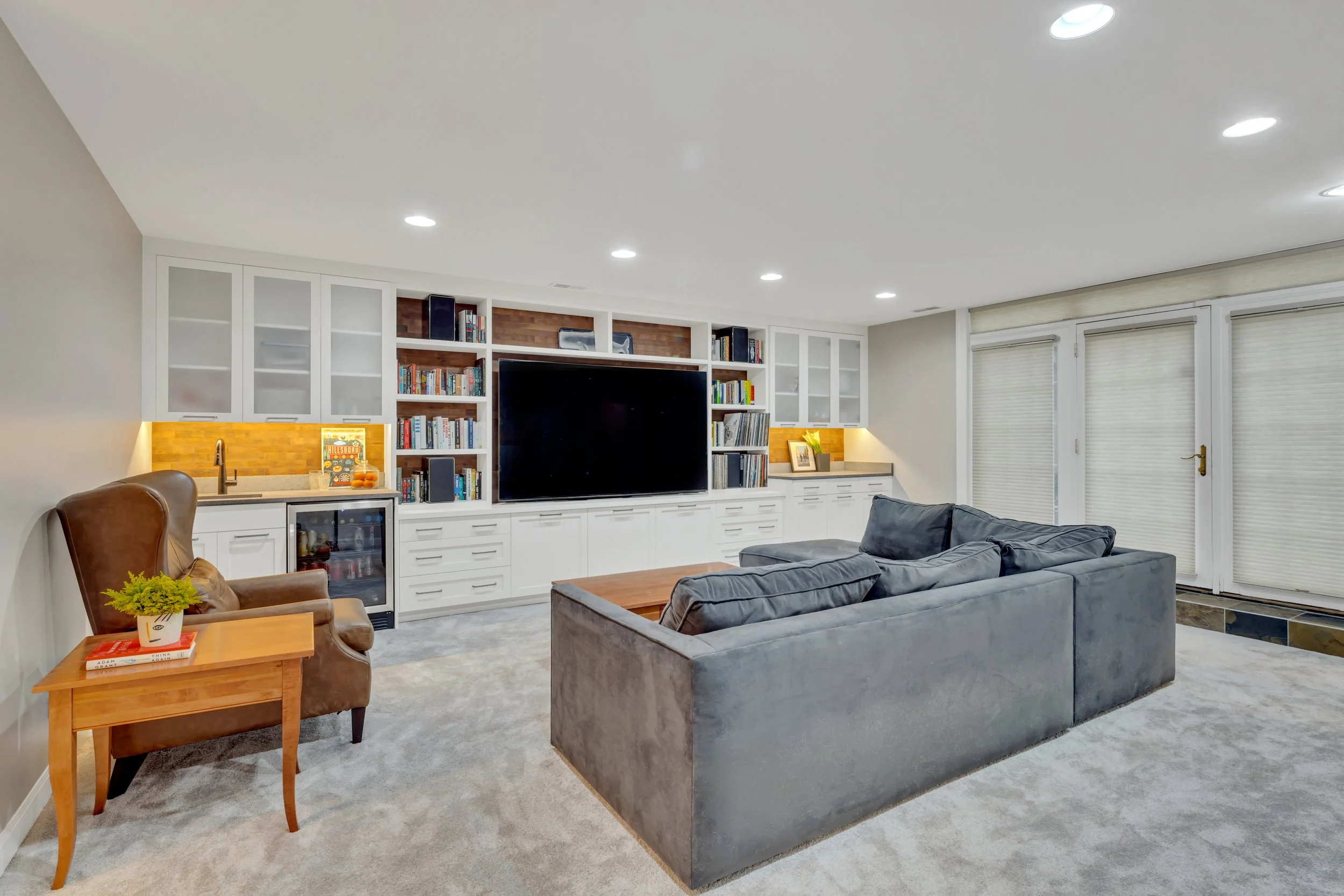 Comfortable living room with a gray sectional sofa, wooden coffee table, and built-in white bookshelves with a large flat-screen TV, books, and decorative items. There's a brown armchair with a small wooden side table and a plant, three windows with 