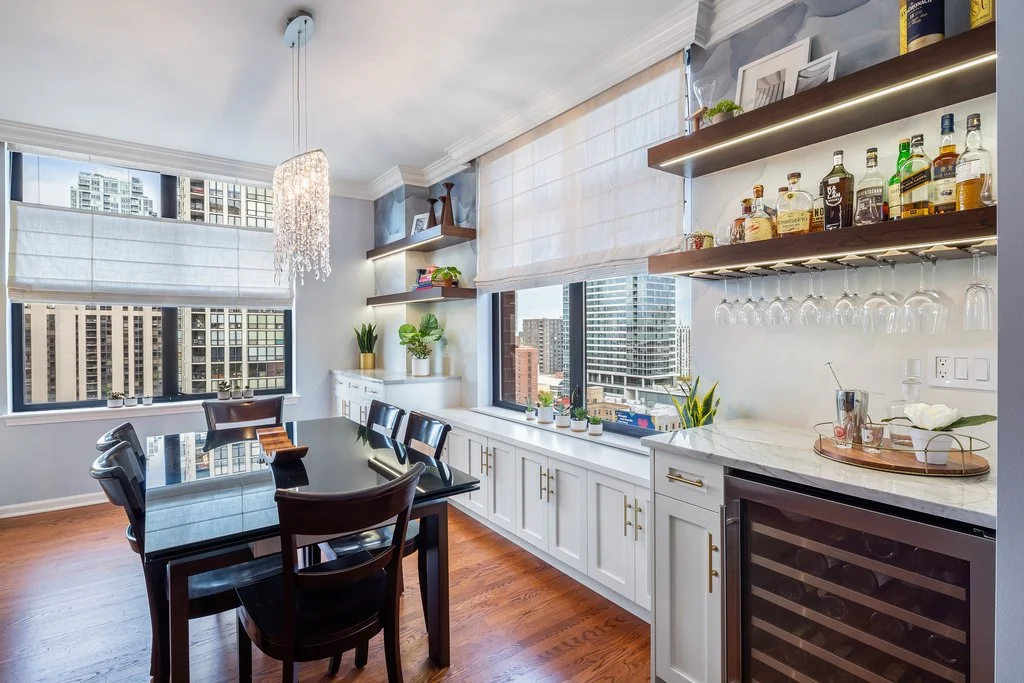 Modern dining area with large windows, a black dining table with six chairs, white cabinetry, shelves with plants and bottles, a wine cooler, and city skyline view.