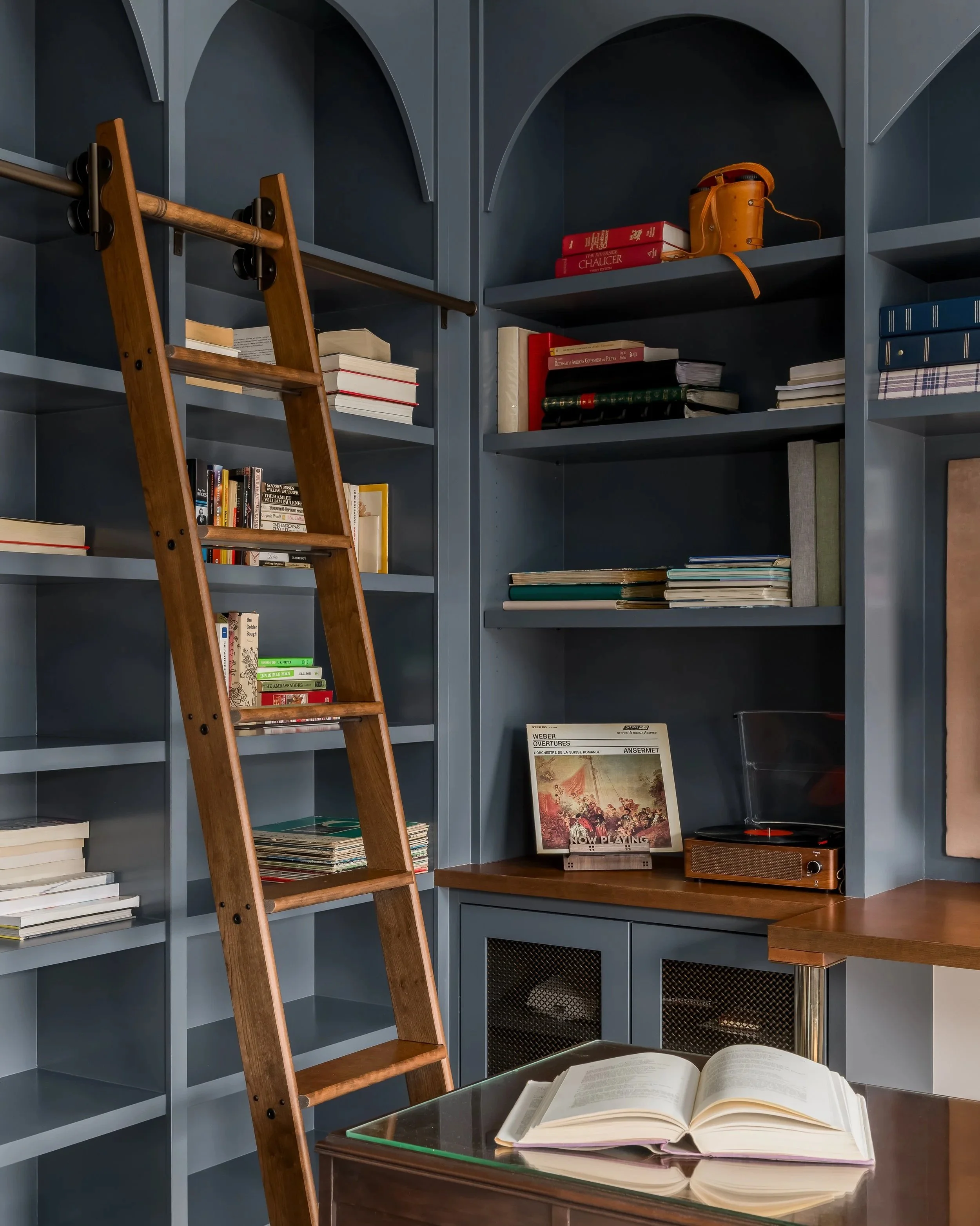 A cozy library corner with a blue bookshelf, a wooden ladder, an open book on a glass table, and a record player.