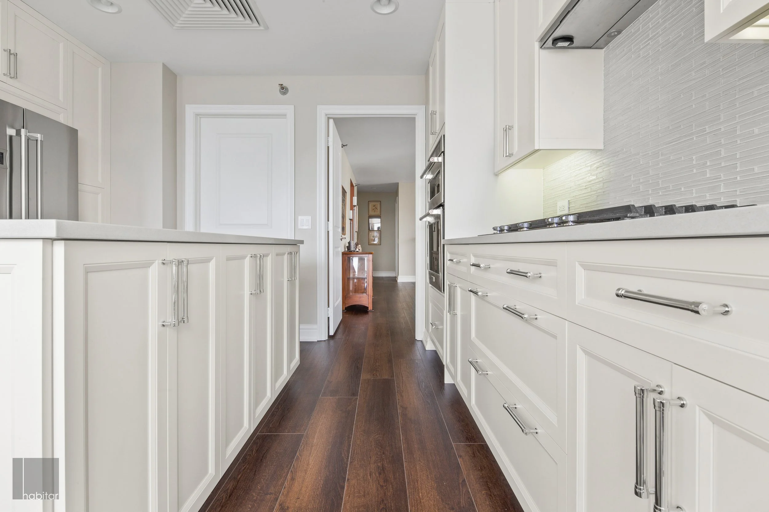 A modern kitchen with white cabinets, stainless steel appliances, and dark wood flooring, leading to a hallway with a small wooden cabinet.