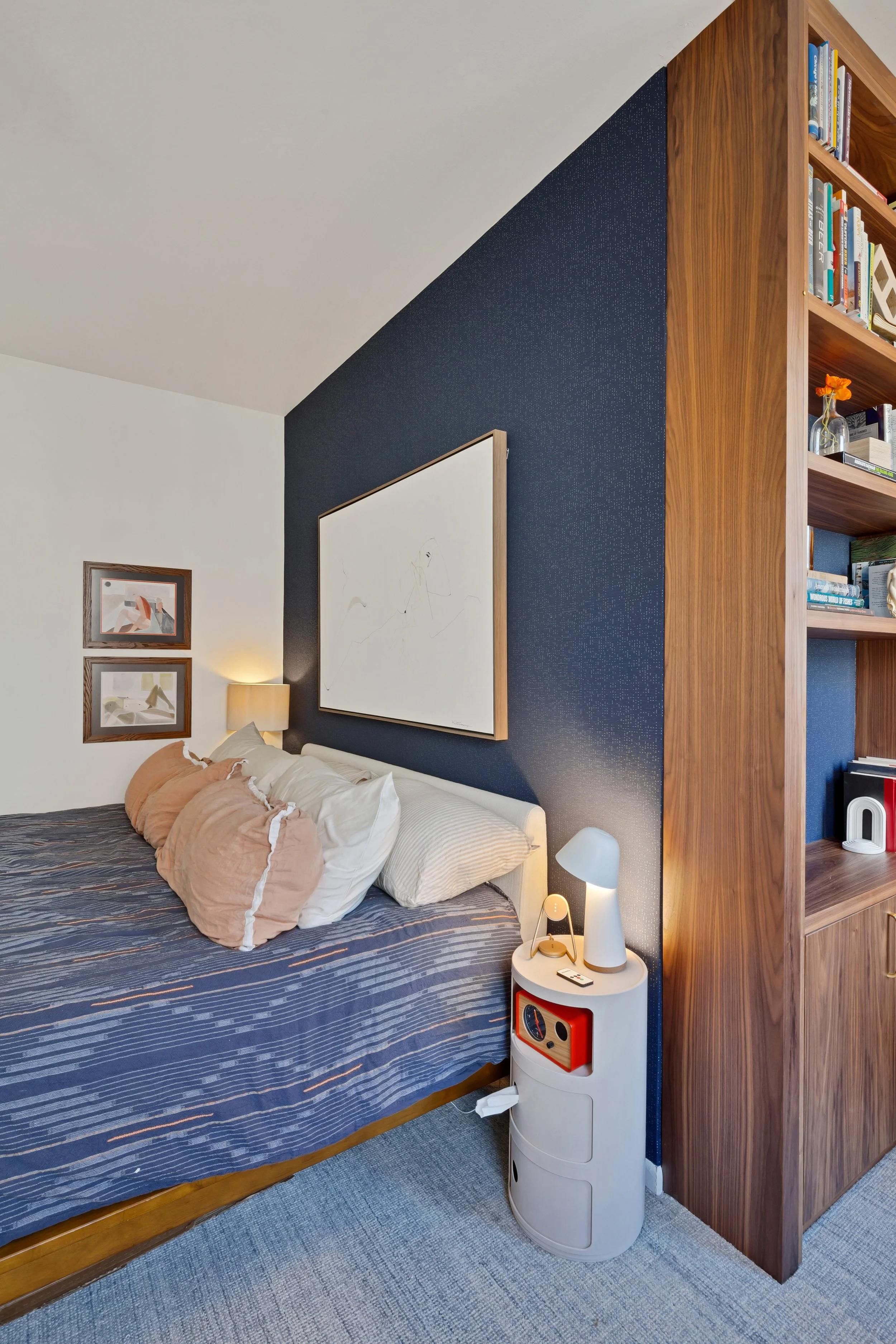 A bedroom with a white bed, beige pillows, and artwork on the wall. A wooden bookshelf is adjacent to the bed. There is a white side table with a lamp and electronic devices. The wall behind the bed is painted dark blue, and the floor is carpeted.