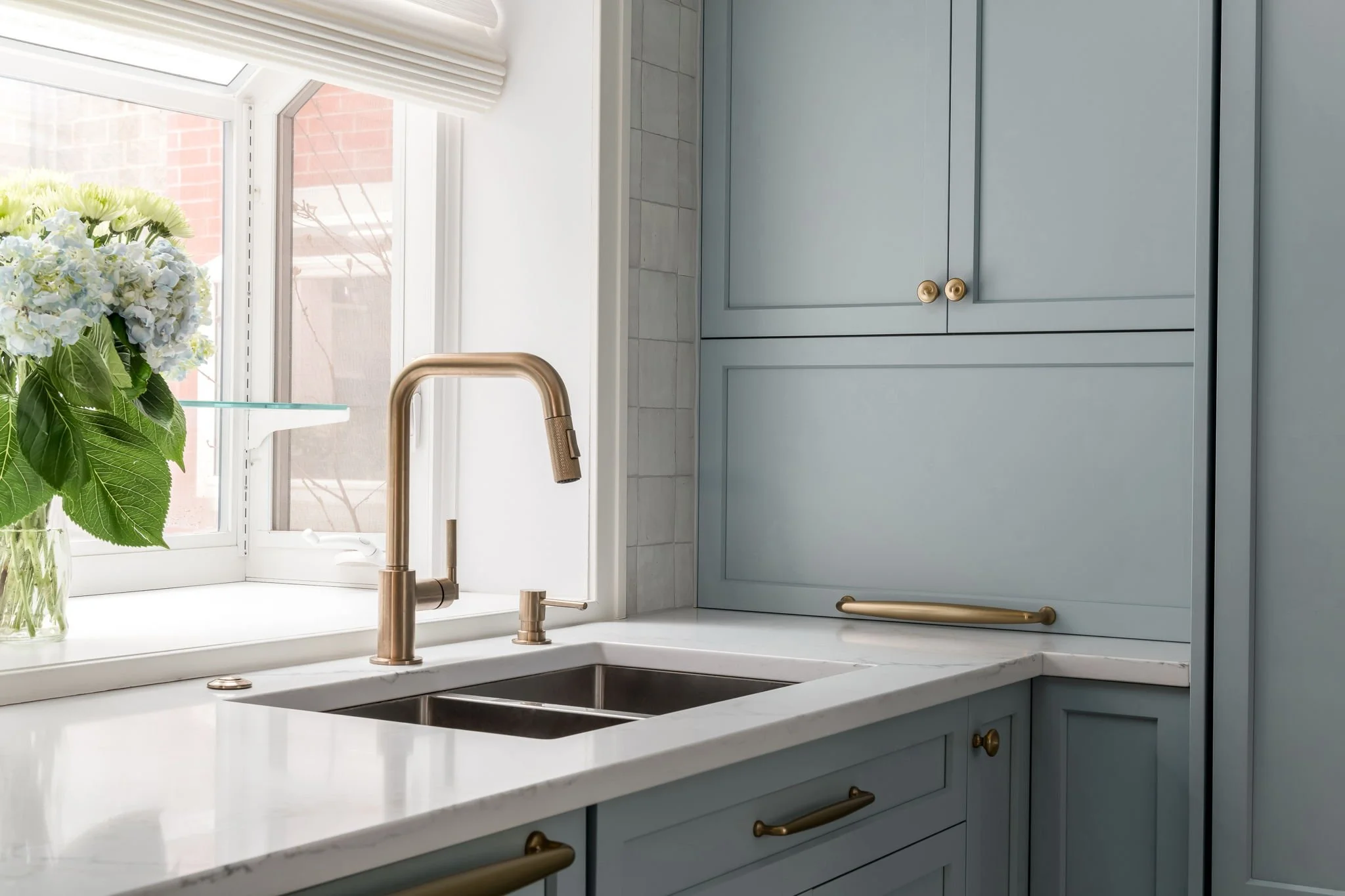 Light blue kitchen cabinet with brass handles, white quartz countertop, and a brass kitchen faucet next to a window with hydrangeas.