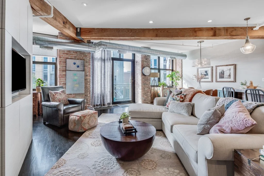 Living room with white sectional sofa, black leather armchair, round wooden coffee table, exposed brick walls, large windows, and modern decor.