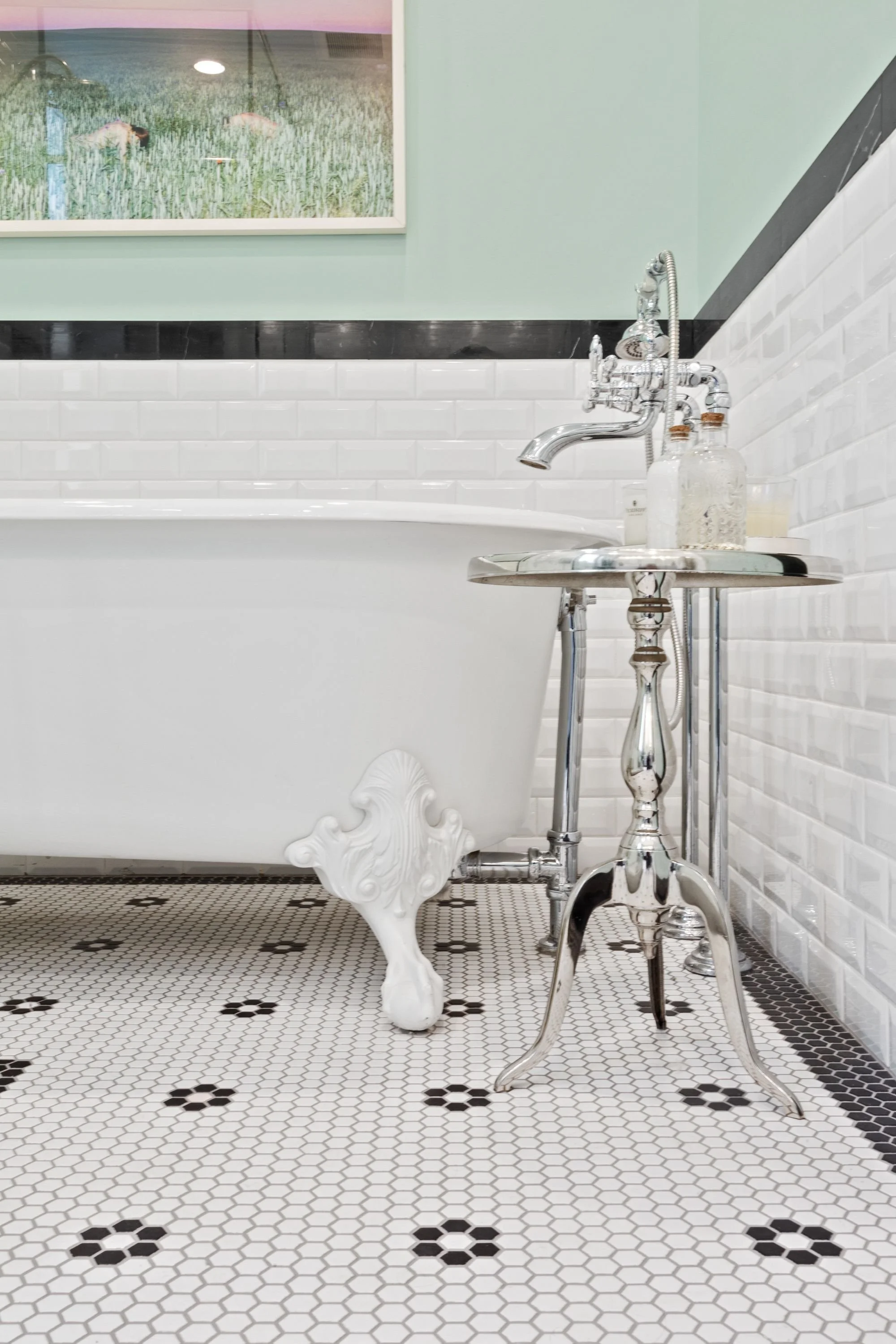 Bathroom with clawfoot bathtub, metallic side table, bottles, and a framed picture of a field of wheat on the wall.