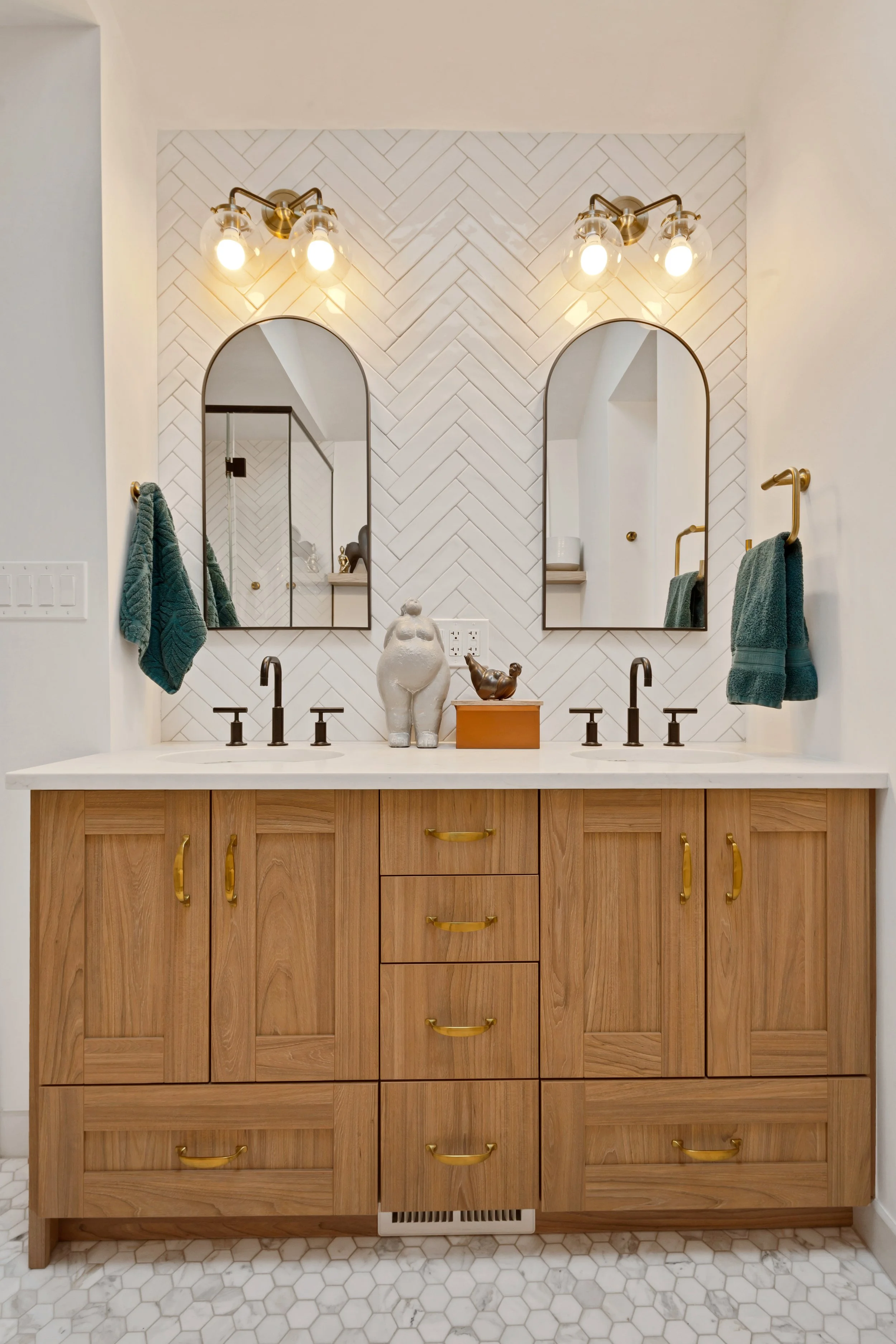Double bathroom vanity with wooden cabinets, white countertop, two black faucets, two mirrors, gold hardware, and teal towels. Decorative figurines and a box are on the countertop. Hexagonal tile floor.
