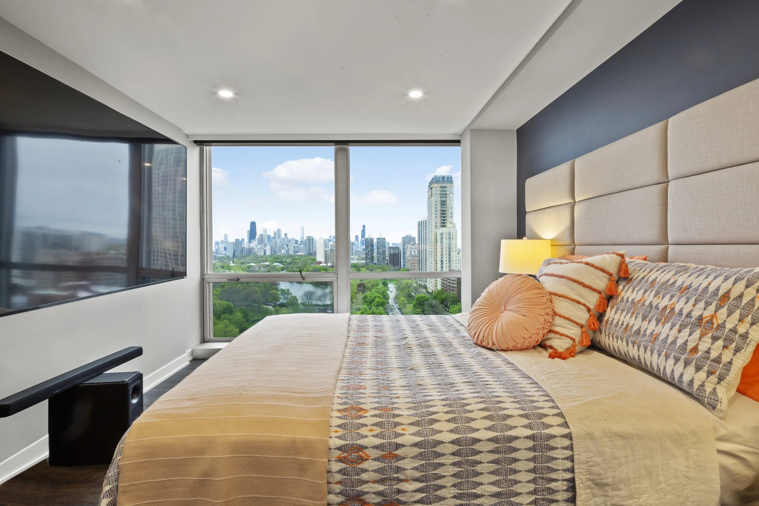 Modern bedroom with large window view of city skyline and greenery, beige upholstered headboard, nightstand with lamp, patterned pillows and bedding.