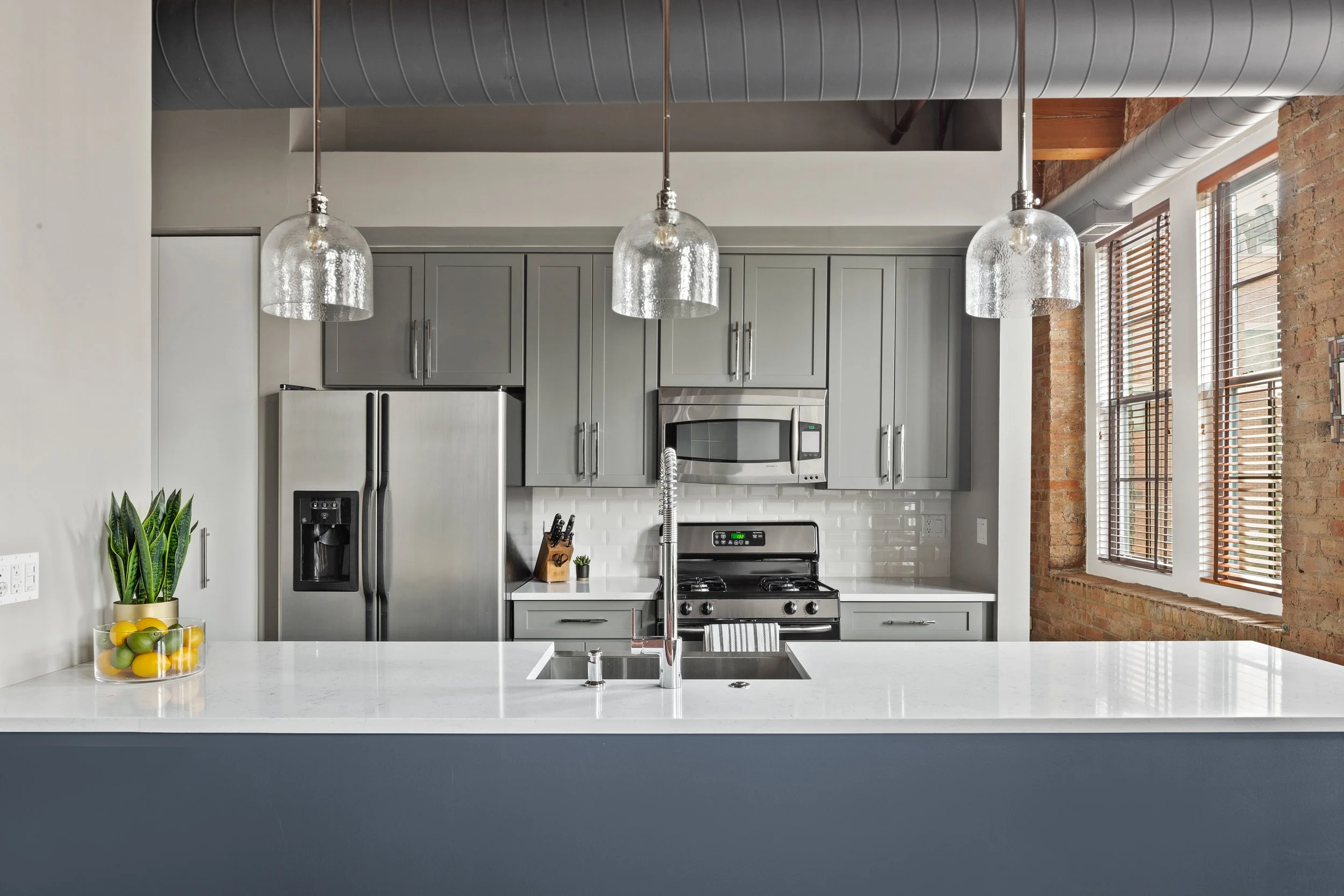 Modern kitchen with gray cabinets, stainless steel appliances, white countertop, and brick walls, featuring pendant lighting and large windows with blinds.