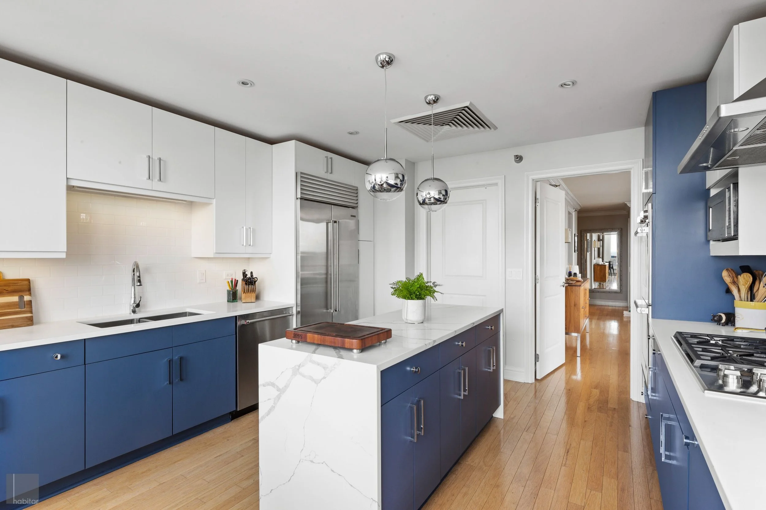 Modern kitchen with blue and white cabinets, wooden flooring, island with a potted plant, pendant lights, stainless steel refrigerator, and a view into a hallway.