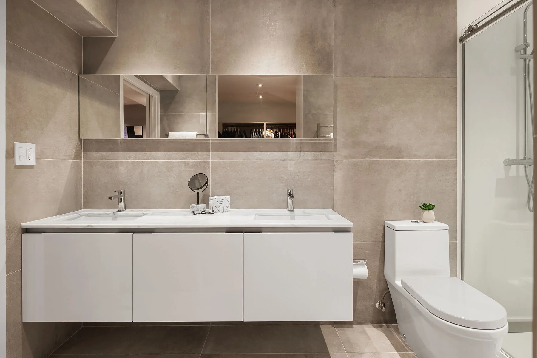 Modern bathroom with a white double sink vanity, large wall mirror, toilet with small potted plant, and a glass shower enclosure.